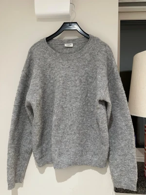 Grå stickad tröja från Kappahl - Mysig grå stickad tröja från Kappahl med rund halsringning och ribbade muddar vid ärmslut och nederkant. Tröjan har en enkel och klassisk design som passar perfekt till kalla dagar. Materialet känns mjukt och behagligt mot huden. Ganska nopprig men det går att få bort.