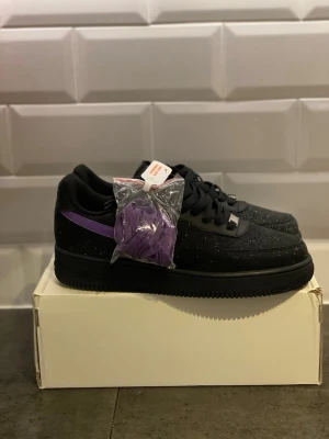 Nike Air Force 1 svart/lila sneakers - Nike Air Force 1 sneakers i svart mocka med vita stänk och en lila Swoosh på sidan. Klassisk låg modell med svart sula och svarta skosnören, extra lila skosnören medföljer. Perfekta för dig som vill sticka ut med en unik färgkombination.
