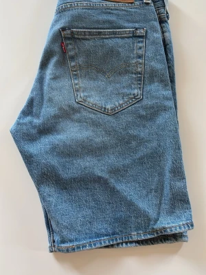 Blå levis jeansshorts - Klassiska blå jeansshorts från Levi's med raka ben och femficksmodell. Shortsen har normal midja, bälteshällor och den ikoniska röda Levi's-lappen på bakfickan. Perfekta för sommaren och tillverkade i slitstark bomullsdenim.