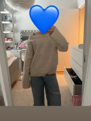 Beige off shoulder stickad tröja - Mysig beige stickad tröja med off shoulder-design och bred ribbad kant vid ärmslut och nederkant. Tröjan har en avslappnad passform och långa ärmar, perfekt för lager på lager och chill stil.