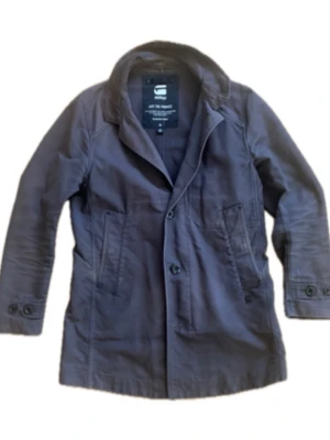 Mörkbrun rock från G-Star RAW - Stilren mörkbrun rock från G-Star RAW med klassisk krage, knäppning framtill och två stora fickor. Tillverkad i ett slitstarkt bomullsmaterial som ger en snygg och avslappnad look. Perfekt för dig som vill ha en tidlös och enkel rock. Saknar två knappar där fram så säljs billigt! OBS. Tveka inte på att höra av er vid frågor!