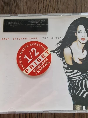 Dana International The Album CD - CD-album med Dana International, släppt av Helicon Records. Omslaget är vitt med en bild på artisten i svartvitt mönstrad klänning. Albumet innehåller låtar som 'Cinque Milla', 'Maganuna' och 'Good Night Europe'. Plastfodral med röd reasticker på framsidan.