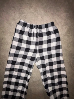 Rutiga pyjamasbyxor i svartvitt - Säljer ett par pyjamasbyxor med svartvitt rutmönster. Byxorna har resår i midjan och dragsko för skön passform samt resår vid bensluten. Perfekta för mysiga hemmakvällar. Materialet känns mjukt och bekvämt. 