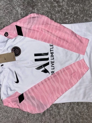 PSG träningströja Nike vit/rosa - Säljer en PSG träningströja från Nike i vitt med rosa mönstrade ärmar. Tröjan har halv dragkedja, svart Nike-logga och PSG-märke på bröstet samt svart tryck med texten 'ALL'. Tillverkad i ett lätt och funktionellt material.