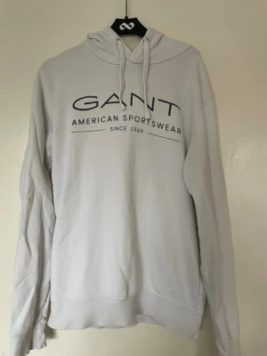Vit hoodie från GANT med tryck - Snygg vit hoodie från GANT med svart tryck på bröstet där det står 'GANT American Sportswear Since 1949'. Hoodien har huva med dragsko och långa ärmar. Tillverkad i mjukt bomullsmaterial som känns skönt mot huden. Perfekt för en avslappnad och stilren look.