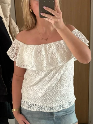 Vit offshoulder spetsblus - Säljer en superfin vit offshoulder blus i spets med volangdetalj över bysten. Perfekt till jeans eller kjol för en söt och trendig look. Blusen har ett vackert spetsmönster och är ärmlös med axelbandslös design.