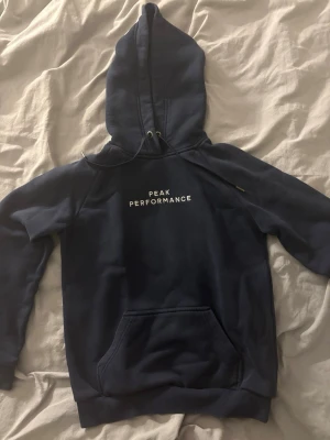 Mörkblå hoodie från Peak Performance - Snygg mörkblå hoodie från Peak Performance med vit logga på bröstet. Klassisk känguruficka framtill, ribbade muddar och justerbar huva med dragsko. Perfekt för en avslappnad och sportig stil.