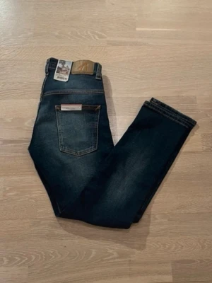 Mörkblå jeans från Nudie  - Säljer ett par mörkblå jeans från Nudie Jeans med klassisk femficksdesign och snygga slitningar. Jeansen har raka ben och bruna kontrastsömmar samt en läderpatch bak i midjan. Perfekta för dig som gillar stilrena och tidlösa jeans.