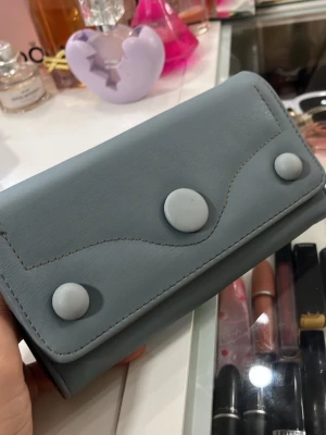 Grå plånbok med knappar - Snygg grå plånbok i konstläder med tre stora dekorativa knappar på framsidan. Plånboken har flera fack inuti, inklusive ett med dragkedja för mynt. Perfekt för att organisera kort och kontanter. Fick den i present aldrig använt 