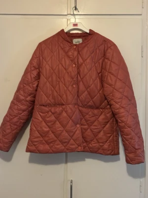 Rostfärgad quiltad pufferjacka Wala - Säljer en snygg rostfärgad pufferjacka från Wala med quiltat mönster och tryckknappar framtill. Jackan har rundad krage och två stora fickor framtill. Perfekt för kalla dagar och ger en trendig look.