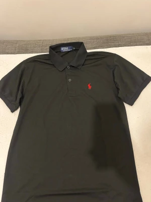 Svart polotopp från Polo Ralph Lauren - Klassisk svart polotopp från Polo Ralph Lauren med korta ärmar och krage. Toppens ikoniska röda broderade logga på bröstet ger en snygg detalj. Materialet är mjukt och känns skönt mot huden. Perfekt för dig som gillar stilrena plagg med en sportig touch.
