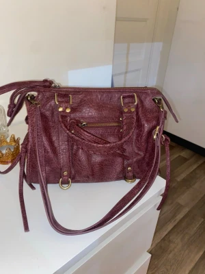 Vinröd handväska med gulddetaljer - Snygg vinröd handväska i konstläder med flätat handtag, justerbar axelrem och guldfärgade metalldetaljer. Väskan har en liten ficka med dragkedja framtill och dekorativa remmar på sidorna. Perfekt för dig som gillar trendiga accessoarer. Använd 2-3 gånger och inga defekter!pris kan diskuteras