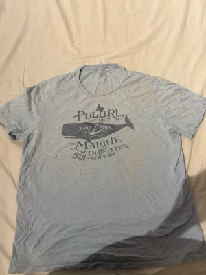 Ljusblå Polo Ralph Lauren t-shirt - Säljer en ljusblå t-shirt från Polo Ralph Lauren med marinblått tryck framtill. T-shirten har klassisk passform, rund halsringning och korta ärmar. Materialet är mjuk bomull, perfekt för en avslappnad stil.