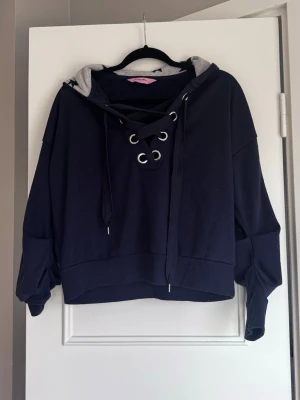 Mörkblå hoodie med snörning - Mörkblå hoodie med breda snören och stora metallöljetter framtill. Huvan är fodrad med grått tyg och blåa stjärnor. 