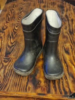 Svarta gummistövlar - Välanvända och bara avspolade, inte ordentligt rengjorda. Innertyget har börjat lossna upptill och det finns två märken på vänsterskon. Ett framme vid tårna och ett långt bak på vänster sida. Stl 27