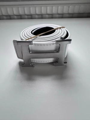 Vitt skärp med stor H-spänne - Snyggt vitt bälte med ett stort, silvrigt H-format spänne. Perfekt accessoar för att lyfta vilken outfit som helst. Gjort i skinn med stilrena sömmar och en clean look. Passar dig som gillar statement-detaljer.