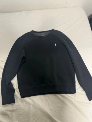 Svart sweatshirt från Polo Ralph Lauren - Klassisk svart sweatshirt från Polo Ralph Lauren med rund halsringning och den ikoniska vita logon broderad på bröstet. Tröjan har ribbade muddar vid ärmslut och nederkant, perfekt för en clean och stilren look.