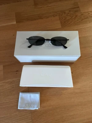 Svarta ovala solglasögon från GAU - Snygga svarta solglasögon från GAU med smal, oval form och minimalistisk design. Bågarna är tunna och svarta, och glasen är mörka. Kommer med vit hård förvaringsbox och putsduk med GAU-logga. Perfekta för en clean och modern stil. Original pris: 800kr.