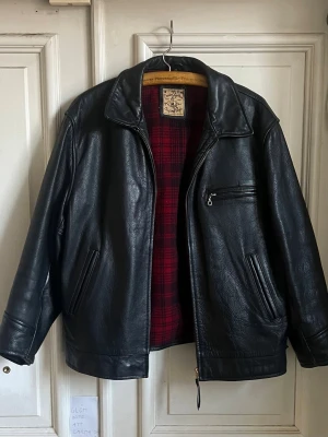 Svart skinnjacka med rödrutig lining - Säljer en klassisk svart skinnjacka från Schott med rödrutig innerfoder. Jackan har dragkedja framtill, tre fickor med dragkedja och bred krage. Perfekt oversized passform och tidlös look som funkar till allt.