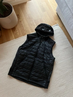 Svart dunväst med huva från C.P. Company - Stilren svart dunväst från C.P. Company med quiltat mönster och två fickor framtill. Västen har en ikonisk huva med inbyggda glasögon och dragkedja hela vägen upp. Perfekt för lager-på-lager och streetwear. Storlek 48 med andra ord M/L