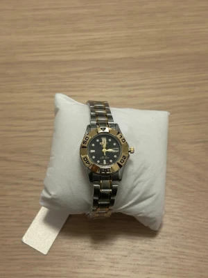 Seiko klocka i silver och guld - Stilren Seiko klocka med länkarmband i silver och gulddetaljer. Svart urtavla med tydliga markeringar och guldfärgade visare. Boetten har en sportig look med guldfärgad ring och robust design. Klockan är inte använt och har fortfarande plast runt armbandet. Klockan väger 55 gram 