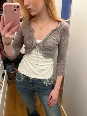 Grå kort bolero med knytning - Supersöt grå stickad bolero med trekvartsärm och knytning framtill. Fint hålmönster och mjukt material som passar perfekt över linne eller klänning. Ger en trendig Y2K-vibe till din outfit.