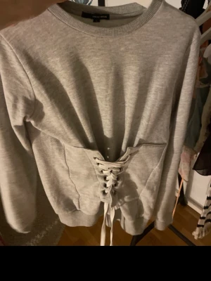 Grå sweatshirt med snörning i midjan - Trendig grå sweatshirt med snörning i midjan som ger en snygg siluett. Tröjan har rund halsringning och långa ärmar. Perfekt för dig som vill ha en basic tröja med en twist.