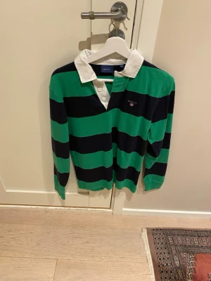Randig rugbytröja från Gant - Snygg långärmad rugbytröja från Gant med breda gröna och mörkblå ränder, vit krage och knappar framtill. Klassisk preppy stil med broderad logga på bröstet. Perfekt för en sportig och avslappnad look.