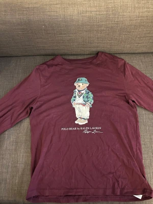 Vinröd långärmad t-shirt Polo Ralph Lauren - Säljer en vinröd långärmad t-shirt från Polo Ralph Lauren med den ikoniska Polo Bear-trycket på bröstet. Tillverkad i mjuk bomull och har en avslappnad passform. Perfekt för dig som gillar klassiska streetwear-vibbar och vill sticka ut lite extra.