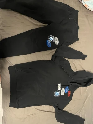 Svart set med patchar från Represent - Säljer ett svart set med hoodie och byxor från Represent. Setet har flera coola patchar och broderade detaljer i vitt, blått och rött på både överdel och nederdel. Hoodien har ficka framtill och byxorna har resår i midjan. Perfekt för en avslappnad och trendig look.