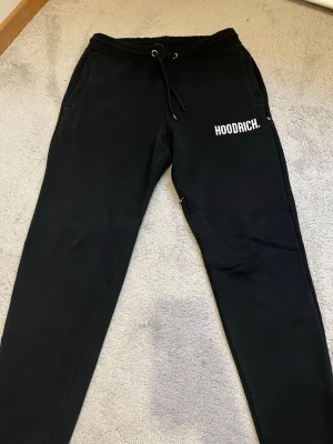 Svarta mjukisbyxor från Hoodrich - Säljer ett par svarta mjukisbyxor från Hoodrich med vit logga på vänster lår. Byxorna har dragsko i midjan, sidofickor och smal passform. Perfekta för chill dagar eller när du vill ha en streetwear-look.