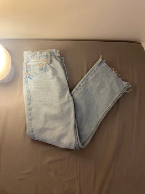 Ljusblå raka jeans med rå kant - Säljer ett par ljusblå jeans med rak passform och rå, avklippt kant nedtill. Klassisk femficksmodell i denim med orange sömmar. Perfekta för en avslappnad och trendig look.