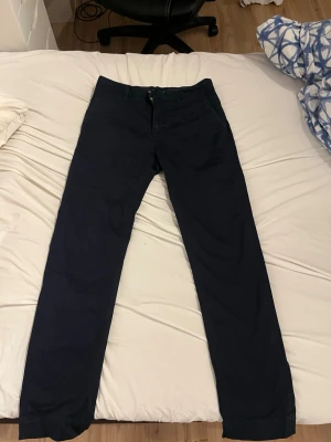 Mörkblå chinos med raka ben från bondelid - Snygga mörkblå chinos med klassisk rak passform och diskreta sidofickor. Byxorna har bälteshällor och två bakfickor med knapp. Perfekta för en stilren och avslappnad look. Materialet känns mjukt och bekvämt, troligen bomull eller bomullsblandning.