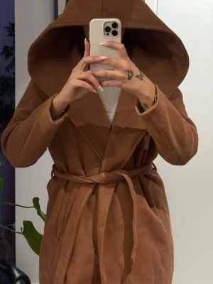Kamelbrun kappa med stor huva - Snygg kamelbrun kappa med stor huva och knytskärp i midjan. Kappan har en avslappnad passform, långa ärmar och två fickor framtill. Tillverkad i mjukt tyg som ger en lyxig känsla. Perfekt för dig som vill ha en trendig och bekväm look. Lite knopprig 