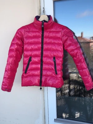 Hot pink dunjacka från Moncler - Snygg rosa dunjacka från Moncler med hög krage och svarta dragkedjor. Jackan har två framfickor med dragkedja och Moncler-logga på ärmen. Tillverkad i glansigt material och fylld med dun, perfekt för kalla vinterdagar.