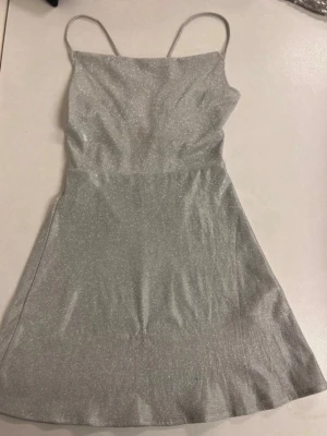 Glittrig silver kortklänning - Supersnygg glittrig silverfärgad kortklänning med tunna axelband och öppen rygg med snörning. Klänningen har en lätt utsvängd kjol och är perfekt för dig som vill sticka ut. Materialet känns mjukt och följsamt, och den glittriga ytan ger en extra wow-effekt. Klänningen har nån blå liten fläck längst ner, men är inte synlig när klänningen är på. 