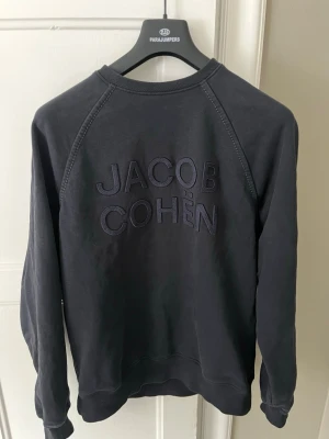 Mörkblå sweatshirt från Jacob Cohën - Snygg mörkblå sweatshirt från Jacob Cohën med broderad logga på ryggen, storlek L passar M. Tröjan har rund halsringning, raglanärmar och ribbade muddar vid ärmslut och nederkant. Perfekt för en avslappnad och stilren look.