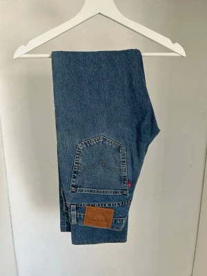Levi's blå raka jeans - Klassiska blå jeans från Levi's i rak modell. Jeansen har en stor bakficka med den ikoniska röda Levi's-lappen och ett brunt lädermärke bak i midjan. Tillverkade i slitstark denim, perfekta för en avslappnad och tidlös stil.