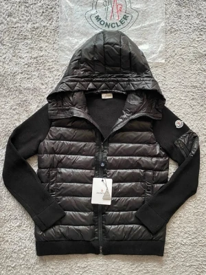 Svart pufferjacka med huva från Moncler - Snygg svart pufferjacka från Moncler med quiltad front och rygg, stickade ärmar och stor huva. Jackan har dragkedja framtill, två sidofickor och Moncler-logga på ärmen. Perfekt för kalla dagar och riktigt trendig look.