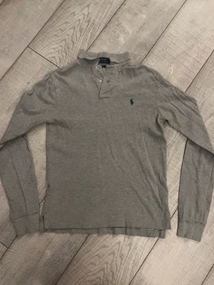 Grå långärmad pikétröja Polo Ralph Lauren - Klassisk grå långärmad pikétröja från Polo Ralph Lauren med krage, knappar vid halsen och den ikoniska blå logotypen broderad på bröstet. Tillverkad i mjuk bomull som är skön att bära. Perfekt till jeans eller chinos.