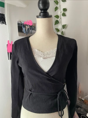 Svart omlottopp med knytband - Svart ribbad omlottopp med långa ärmar och knytband i midjan. Toppen har en djup v-ringning och sitter snyggt figurnära. Perfekt att styla med ett linne under för en trendig lager-på-lager-look.