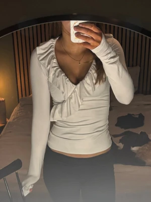 Vit omlottopp med volang - Snygg vit omlottopp med djup v-ringning och volangdetaljer längs halsen och framtill. Långärmad och figurnära passform, perfekt för att lyfta vilken outfit som helst. Tillverkad i mjukt stretchigt material.