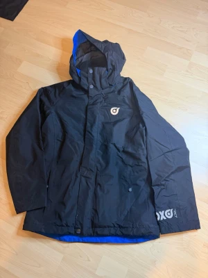 Svart vindjacka från Woxo KappAhl - Svart vindjacka från Woxo by KappAhl med blått foder och huva. Jackan har dragkedja och tryckknappar framtill, samt logga på bröstet och ärmen. Praktisk ficka framtill och justerbar huva. Perfekt för blåsiga dagar och streetwear-stil.