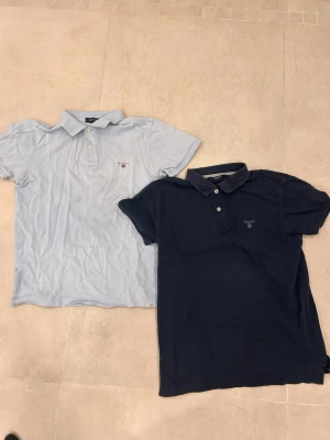 Två pikétröjor från Gant, blå & ljusblå - Säljer två klassiska pikétröjor från Gant, en i marinblått och en i ljusblått. Båda har korta ärmar, krage och knappar framtill samt diskret Gant-logga på bröstet. Perfekta för en clean och stilren look. För dig som är 160-168