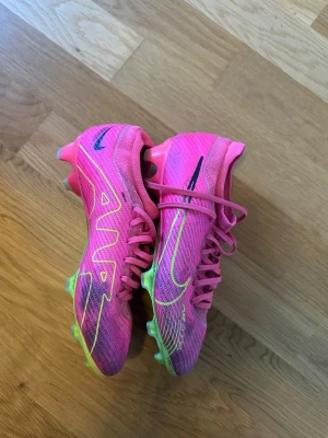 Nike rosa Mercurial fotbollsskor pro - Säljer ett par Nike Mercurial fotbollsskor i en riktigt snygg rosa färg med neon- och svarta detaljer. Skorna har snörning, låg profil och mönstrad ovandel i syntetmaterial. Sulan är delvis neonfärgad och har fasta dobbar för spel på gräs.