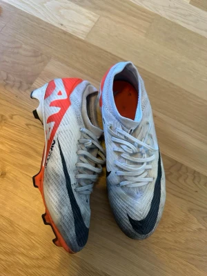 Nike Mercurial vita fotbollsskor pro - Säljer ett par Nike Mercurial fotbollsskor i vitt med svarta och röda detaljer. Skorna har snörning, låg profil och mönstrad sula med fasta dobbar för bra grepp på planen. Ovandelen är i syntetmaterial för lätt känsla och bra bollkontroll.