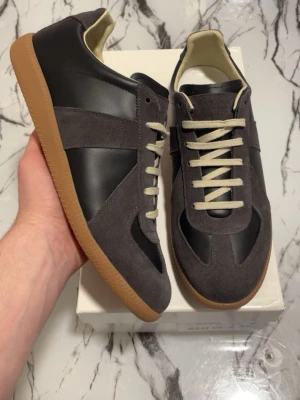 Maison Margiela Replica sneakers grå/svart - Maison Margiela Replica sneakers i grå mocka och svart läder med beigea skosnören och klassisk gummisula. Snygg retrostil med låg profil och vita detaljer på insidan. Perfekta för dig som gillar minimalistisk och stilren design.