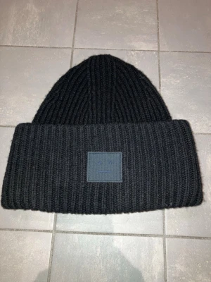 Svart ribbstickad mössa Acne Studios - Säljer en svart mössa från Acne Studios med den klassiska ansiktsloggan framtill. Mössan är i tjockt, mjukt material och har en bred uppvikt kant.❣️
