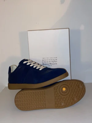 Blå Maison Margiela sneakers - Snygga blå sneakers från Maison Margiela med ovandel i mocka och läder, vita snören och klassisk gummisula i brunt. Skorna har en stilren och tidlös design som passar perfekt till streetwear och casual outfits.