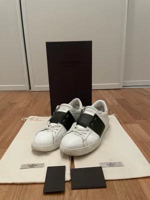 Valentino open i färgen kahki - Säljer ett par snygga vita sneakers från Valentino med ett brett olivgrönt band över mitten. Skorna har klassisk låg profil, vita snören och detaljer i läder. Perfekta för dig som vill ha en stilren och exklusiv look. De har en lite. Defekt på insidan av hälen och är även målade längst fram men inte för att de har varit något fel me de bara pga längre hållbarhet.